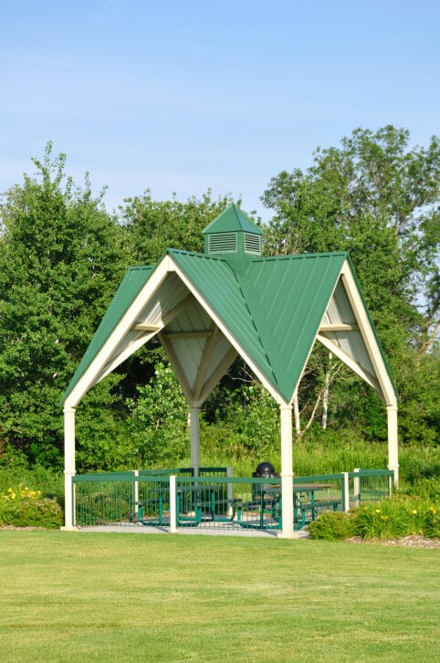 A tall gable roof in a bright green makes this public gazebo an easy structure to spot from a long ways away. The gazebo is furnished with a picnic table, chairs, and a trash can.