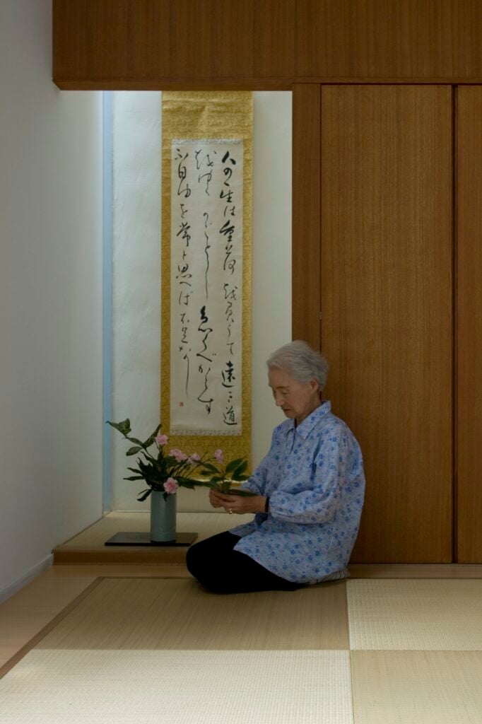 The homeowner is seen practicing ikebana, the art of flower arranging, in a private area of the home. Soft dark natural wood tones on the wall and ceiling stand over light hardwood flooring and more of the bamboo area mat.