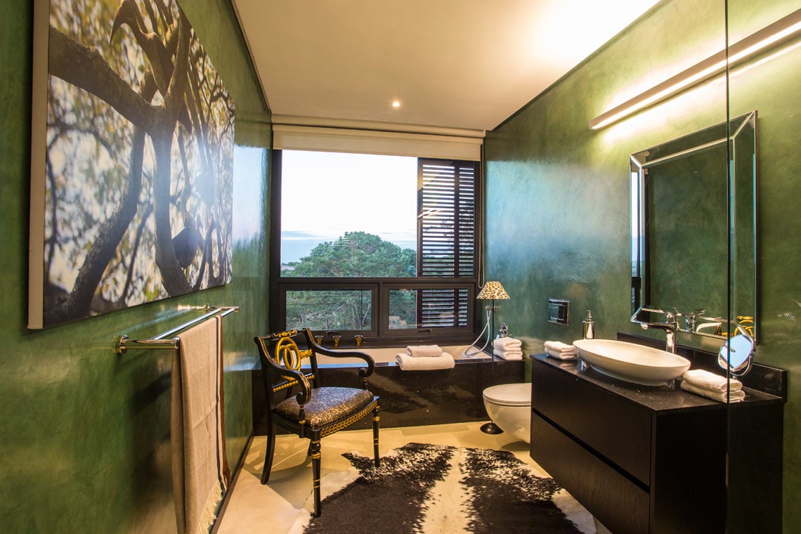 Green primary bathroom designed with a lovely tree wall art canvas. It has a dark wood sink vanity and rectangular tub framed with black marble along with an accent chair.