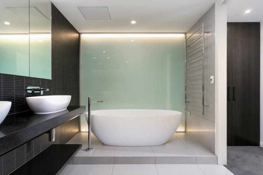 Primary bathroom accented with matte black subway tiles that complement the black marble counter where dual vessel sinks sit. It also has a pedestal tub across a frosted glass window over white tiles flooring.