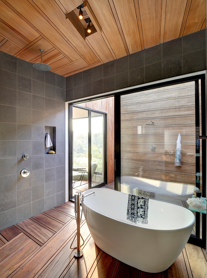Contemporary primary bathroom with gray tiled walls and hardwood flooring. It is enclosed with a black framed glass doors that open to the outdoor rinsing station. 