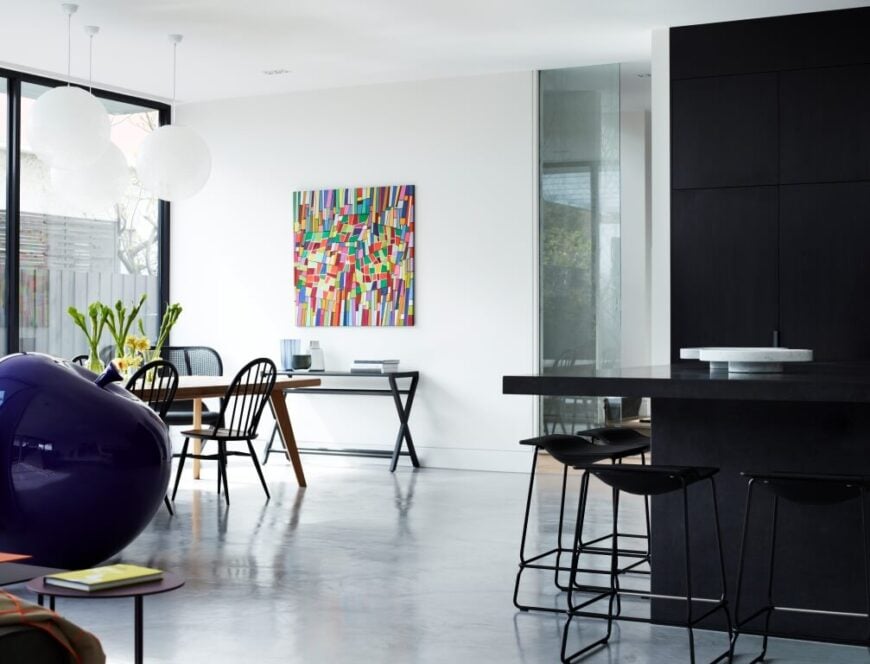 Across the wide expanse of white flooring, we see the dining room area, with natural wood table and black chairs beneath a trio of spherical lights. A fractal rainbow painting hangs on the white wall, adding a small but powerful bloom of color in the room.