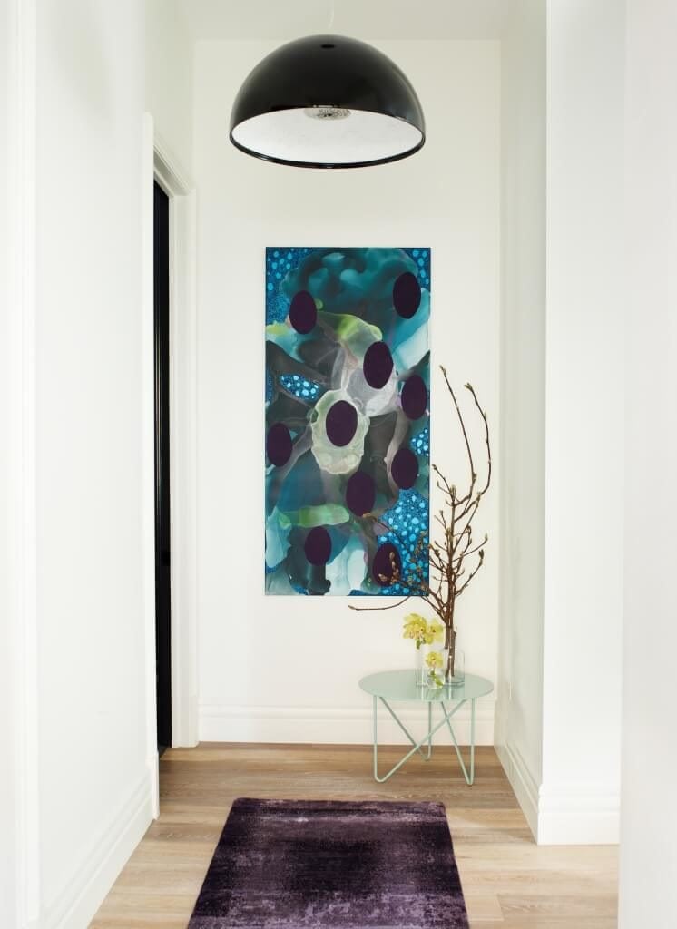 The hallway to the private areas of the home features light hardwood flooring, for a softer pairing with the white walls. Furthering the overall theme, a single colorful painting and purple rug puncture the neutral palette.