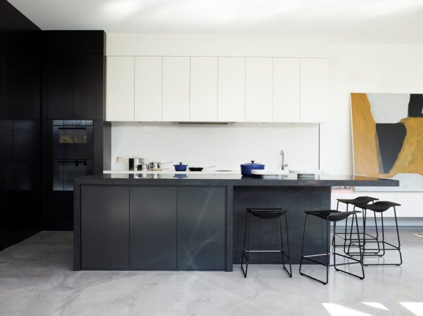 The kitchen centers on a large charcoal toned island, its contrast emphasized by both pristine white and dark wood cabinetry. The island houses an extended countertop, with space for several modern black bar stools.