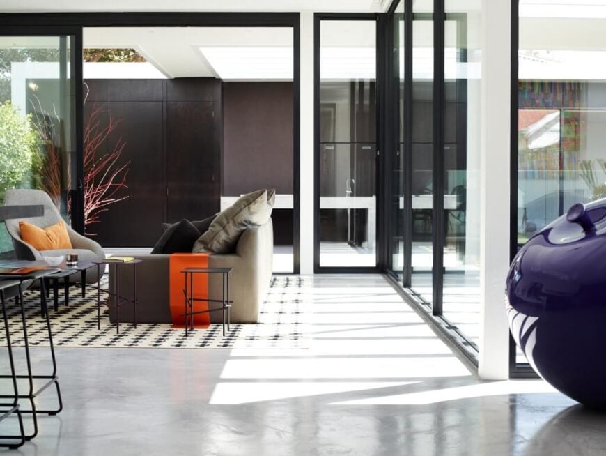 Viewed from the dining area, we see the minimalist living room setup wrapped in glass and, at left, rich dark wood wall panels. Sunlight fills this entire open space, highlighting the spare bursts of color.