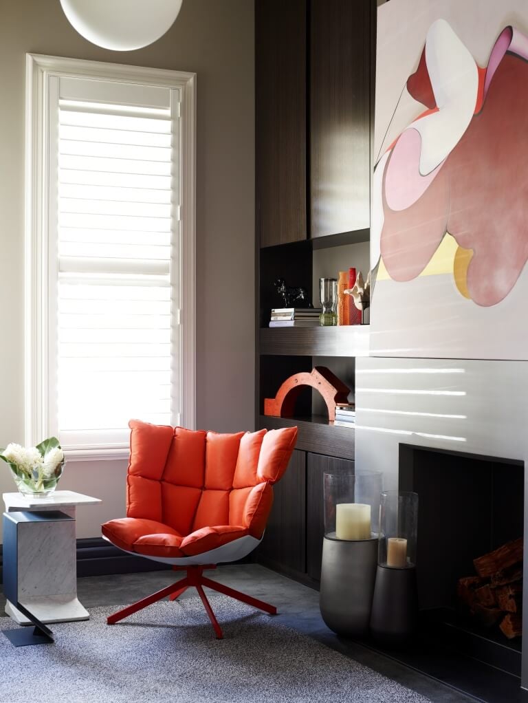 This cozy home office space features another subtle burst of color, with a plush modern orange chair adding a hint of playfulness to the stately, neutral toned room. A fireplace at right is topped by a colorful painting, with rich wood shelving filling out the corner.