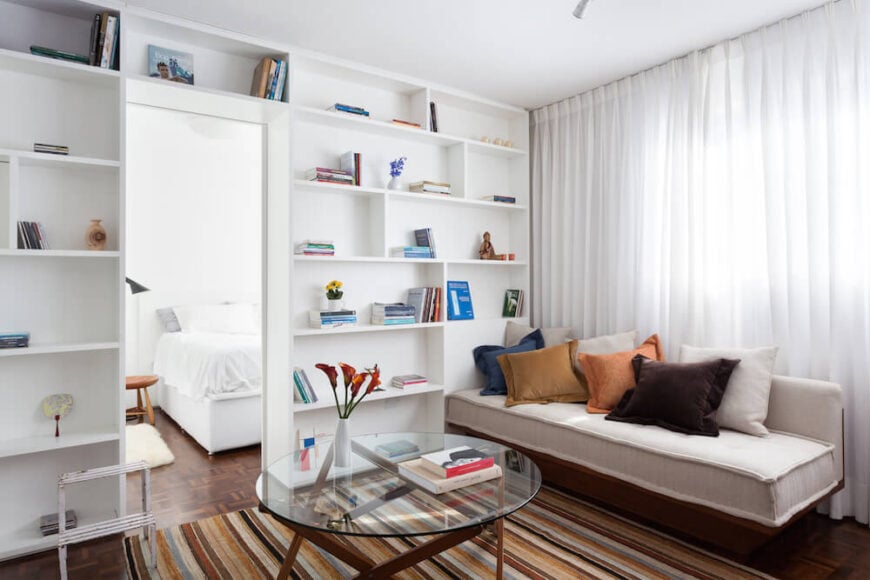 Here, we have a photo of the main part of the house. The living area has a beautiful sheer white curtain that lining the wall and floor-to-ceiling shelving. The hues in the carpet complement the colors of the pillows on the couch for a unified scheme.