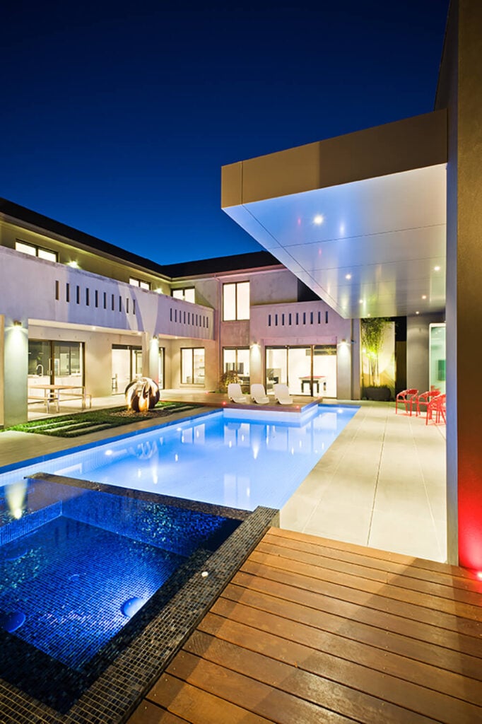 The jacuzzi is defined by its body of dark micro-tiles, distinct from the wood deck and concrete slabs surrounding. Above and to the left, we can see a broad second floor balcony overlooking this area.