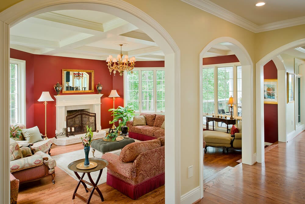 Large formal living room boasting red walls with white accent. The room also offers a cozy set of seats and a fireplace, lighted by a gorgeous chandelier hanging from the coffered ceiling.