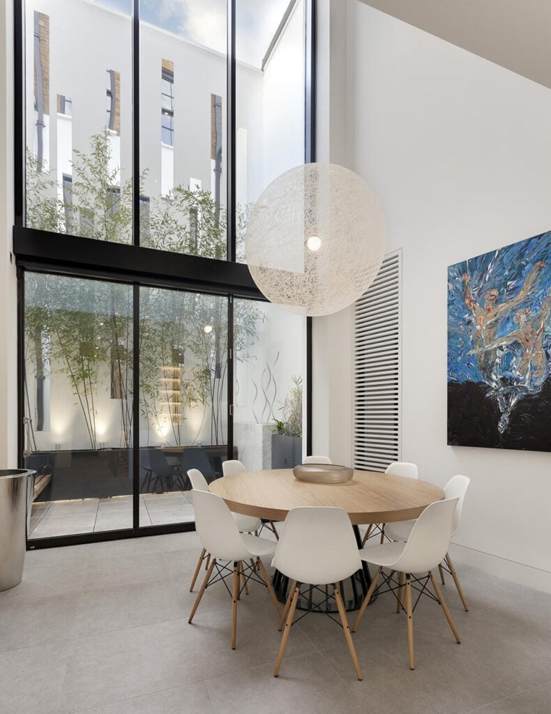 The dining room is wrapped in white and light colors, positively glowing in the natural light provided by the massive, full height windows. A minimalist natural wood table stands surrounded by midcentury modern chairs beneath a large, transparent spherical chandelier.