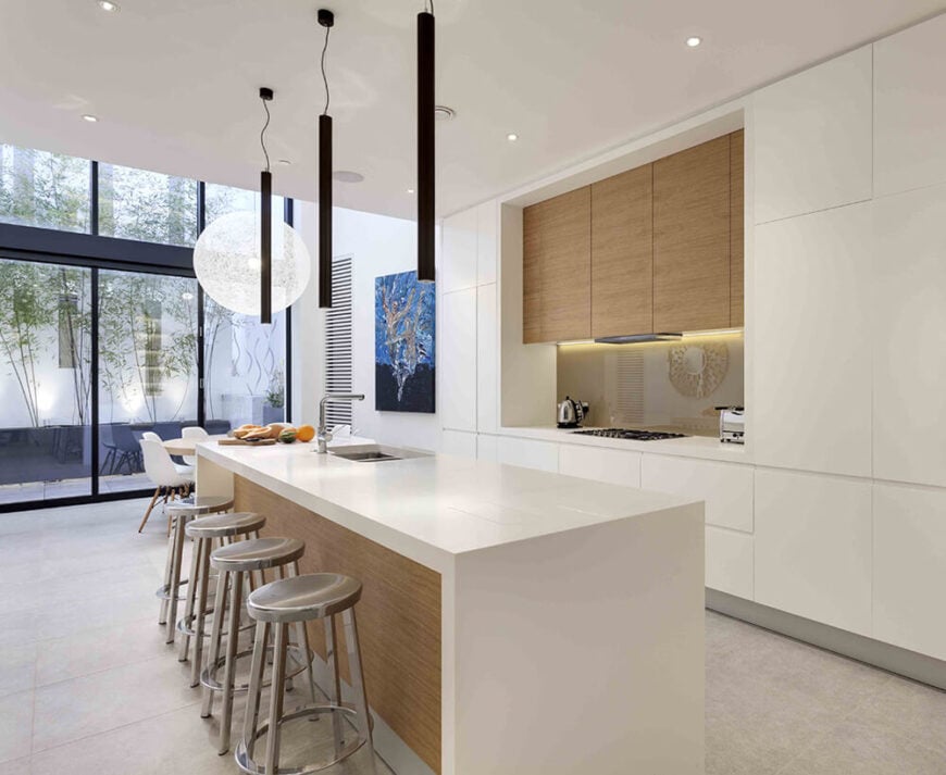 Moving back into the kitchen, we see that the white minimalist space centers on a lengthy island with built-in sink and plenty of room for diners. Light natural wood accents highlight this entire area.