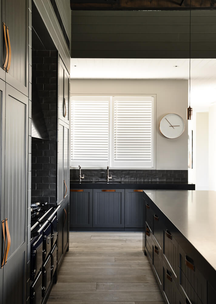 Moving inside the kitchen, we can glimpse the elegant dark toned cabinetry with unique leather strap handles, adding a touch of old school farmhouse charm. The countertops and oven appear in black as well, keeping the tone neat.
