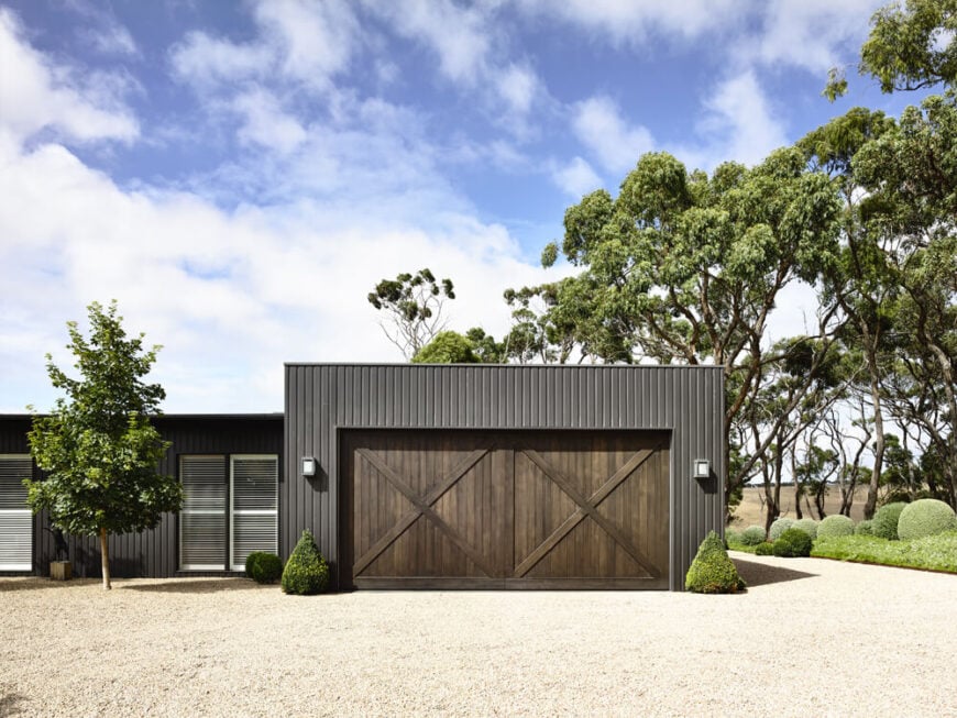 The garage helps maintain that farmhouse look with large cross-beam doors in a dark coffee brown tone. All around, we can see the carefully crafted hedges and sculpted bushes surrounding the home.