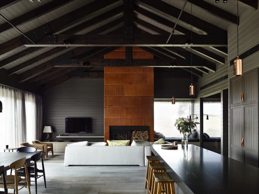 Moving indoors, we come to the large open plan central space, which includes the living room, dining, and kitchen functions. This area centers on the massive rich brown fireplace, towering over the mostly greyscale interior.