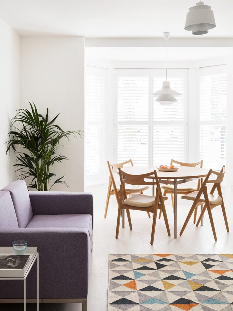 The dining area is part of the larger open plan space at the center of the home. Here we see how elements like the natural wood dining set, contemporary purple sofa, and multicolored rug offer texture and variety in the white space.