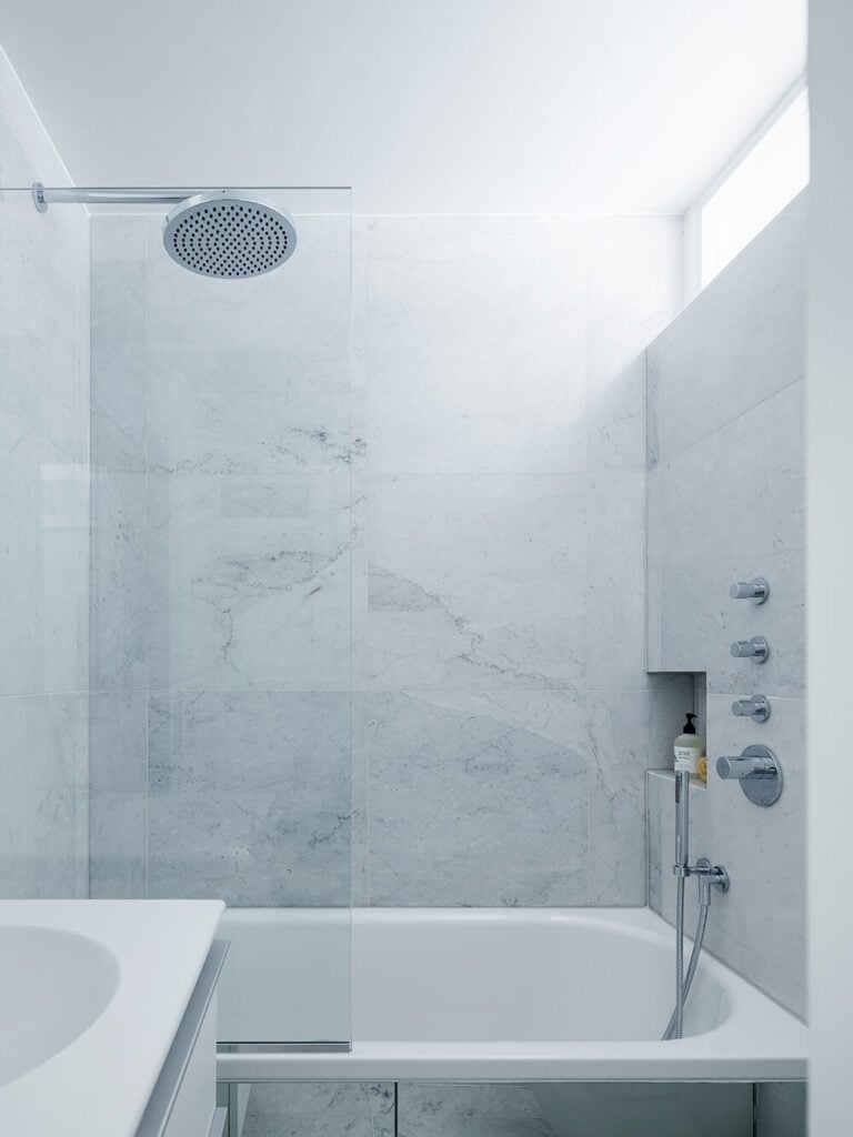 The primary bath is awash in white marble, adding a hearty textural element to the white minimalist look. A large soaking tub is framed in glass, while a slim upper window offers some sunlight to the room.