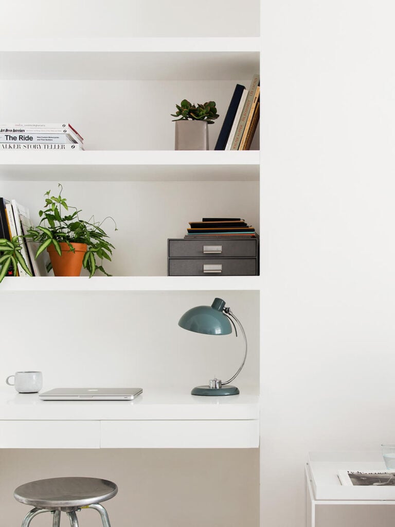 A cozy home office space features a built-in desk and abundant shelving, all in white and meshing perfectly with the walls. Subtle instances of color and chrome spike the minimalist palette.