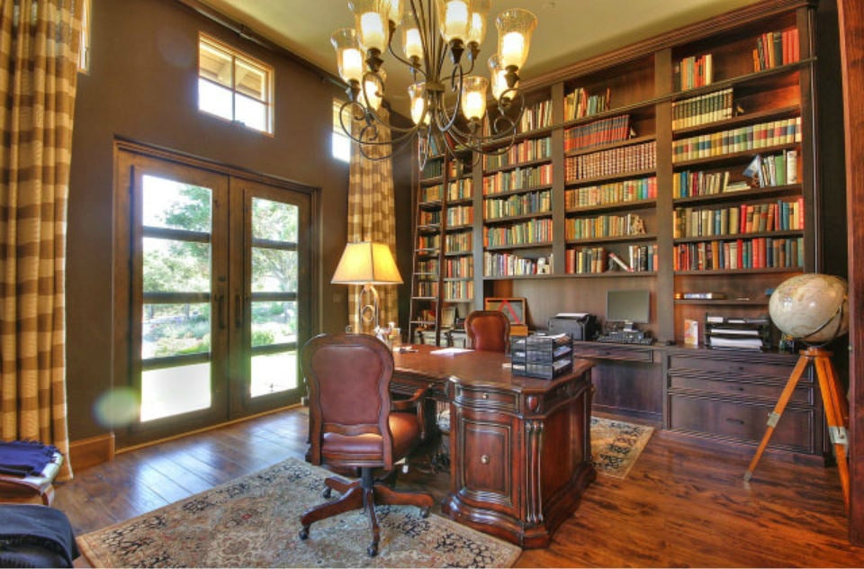 Tuscan home office with a wood plank flooring and a french door covered with striped draperies. It includes a carved wood desk facing the full height bookshelf that's illuminated by a wrought iron chandelier.
