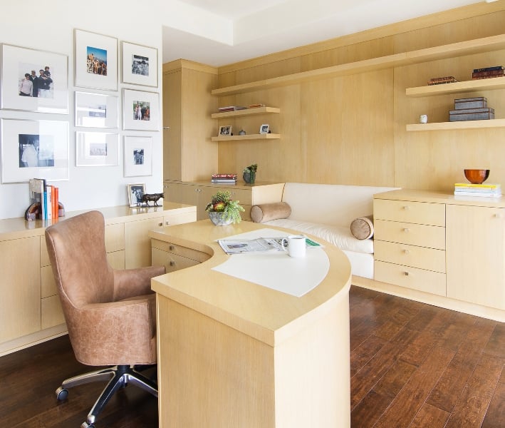 Charming home office features white gallery frames mounted above the light wood cabinet that matches with the curved office desk and built-in storage fitted with a white seating in the middle.