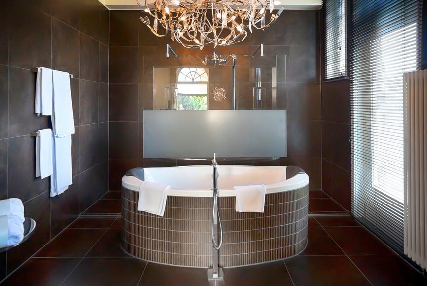 Sophisticated primary bathroom with a fancy chandelier that hung over a circular pedestal soaking tub. It has an open shower area and a glazed window covered with white blinds.