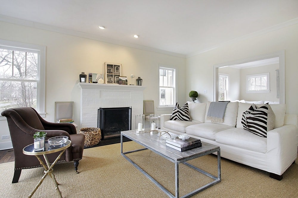 Classy white formal living room with an elegant fireplace and a chanting sofa set on top of the brown rug covering the hardwood flooring.