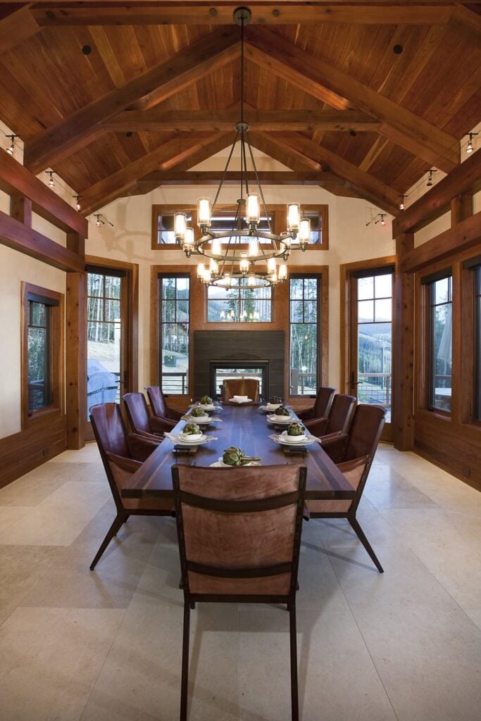 Rustic dining room illuminated by wrought iron chandeliers that hung from a cathedral ceiling with exposed wood beams.  It includes wooden rectangular table with matching dining chairs.