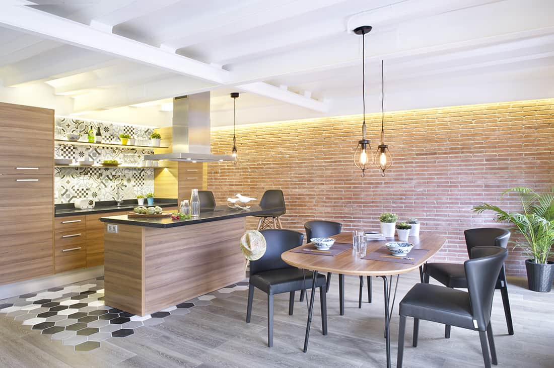 This kitchen area with brick wall and white ceiling with beams provides a nice kitchen counter with a breakfast bar, along with a dining table set lighted by pendant lights.