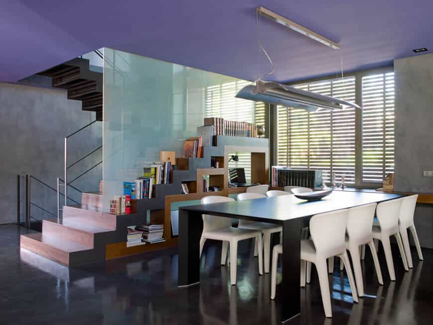 This dining room is situated across the staircase with built-in storage and frosted glass that serves as a divider wall. It includes a black dining table with white chairs over concrete flooring.