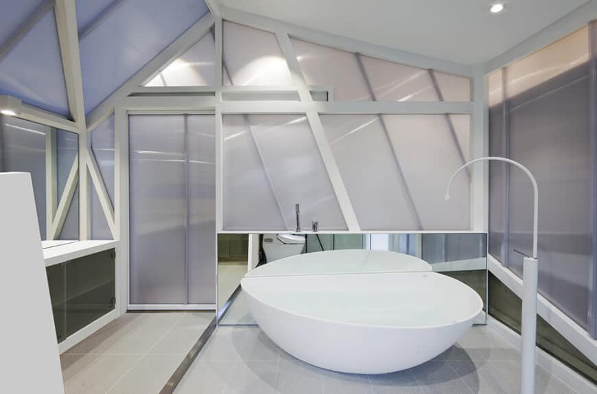 Minimalist primary bathroom surrounded with white framed frosted glass walls along with vaulted ceiling. It has a pedestal tub fixed to the mirror paired with a standalone faucet.