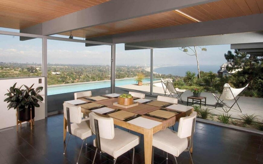 Luxurious glass-walled dining area with a stunning view of the pool and the ocean, with a simple wooden table for six, white upholstered chairs, and dark hardwood floors.