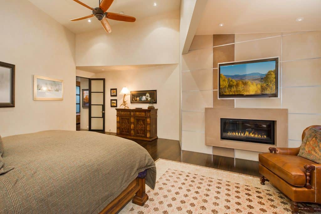 Contemporary primary bedroom with a wooden bed frame and leather accent chair that sits on a shabby chic rug over hardwood flooring. It features a fireplace and display panel mounted on the striped marble wall.