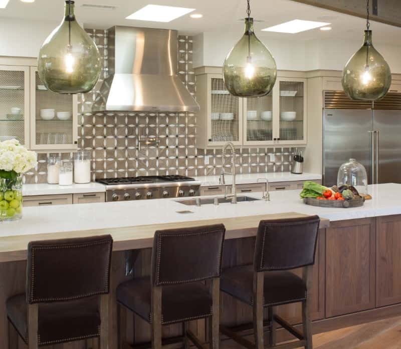 The beautiful and charming green glass hoods of the pendant lights stand out in this Tropical-style kitchen that has simple white countertops paired with a light gray patterned backsplash. These are all illuminated by the skylights of the white ceiling.