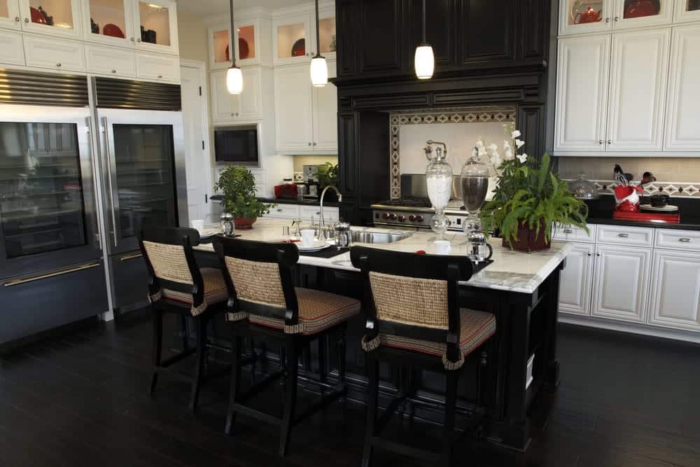 This Tropical-style kitchen has black hardwood flooring that blends with the black wooden kitchen island and the large wooden structure that houses the stove-top oven and its vent. This is flanked by the contrasting white cabinetry of the peninsula that has glass panels on top for the decorative dishware.