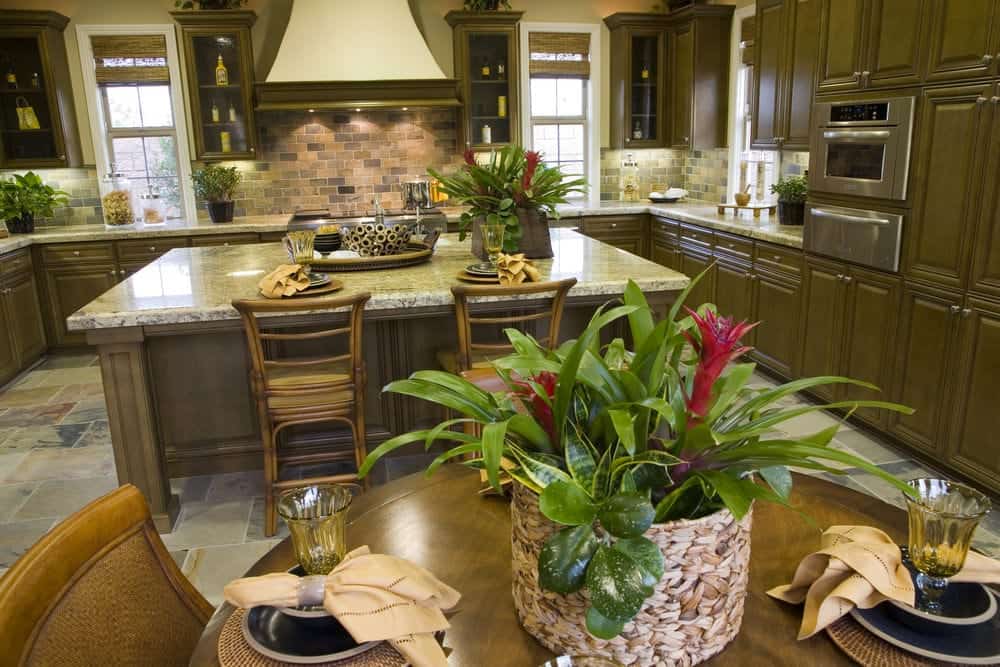 The U-shaped peninsula of the Tropical-style kitchen green wooden cabinets and drawers that matches the various potted plants that adorn the marble countertops. These are then paired with brick backsplash that are lit by the lights underneath the floating cabinets.