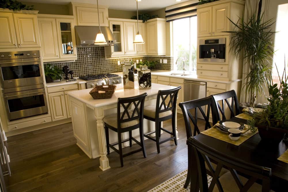 The small space of this Tropical-style kitchen is maximized by the small wooden white kitchen island that matches the L-shaped peninsula and its shaker designs on its cabinets. These white wooden elements serve as a nice background for the potted plants placed on various spots.