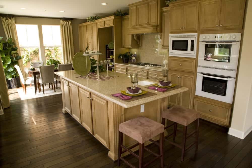The dark hardwood flooring has a homey plank finish to it that complements the wooden cabinetry that has a slightly lighter shade. These are adorned with plants on the space between their top and the white ceiling that has recessed lights complementing the white modern appliances.