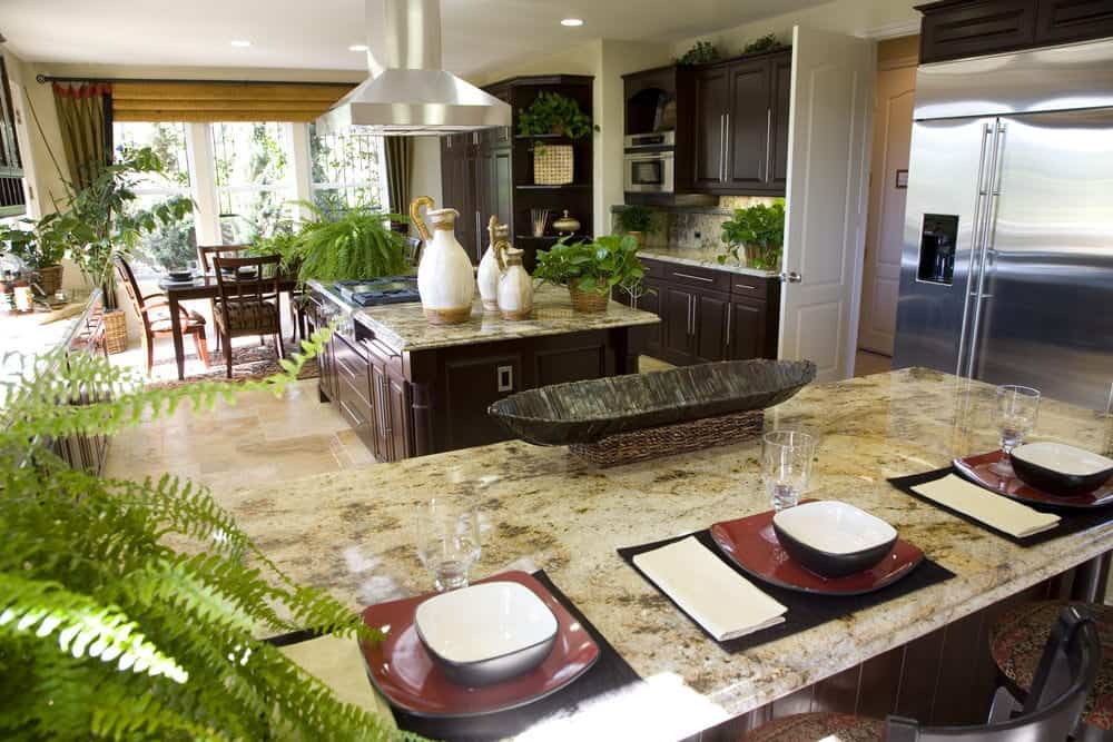 The various ferns and potted plants stand out in this Tropical-style kitchen that has dark brown cabinets and drawers paired with marble countertops that match the beige marble flooring tiles.