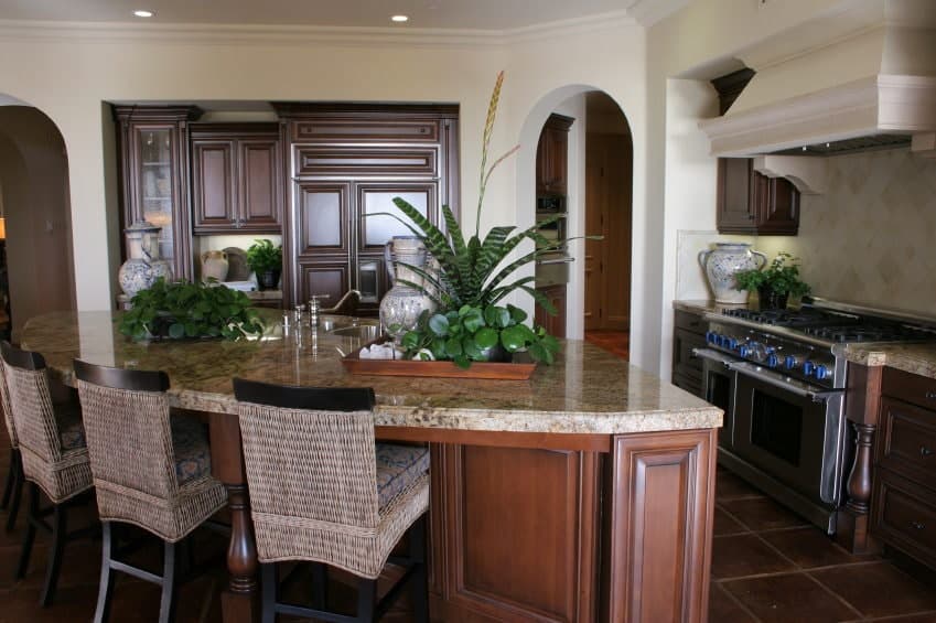 The unique curved wooden kitchen island has a light brown marble countertop that goes well with the terracotta flooring tiles and the woven wicker stools with black legs. All of these dark elements are contrasted by the white walls and ceiling blending with the vent hood.
