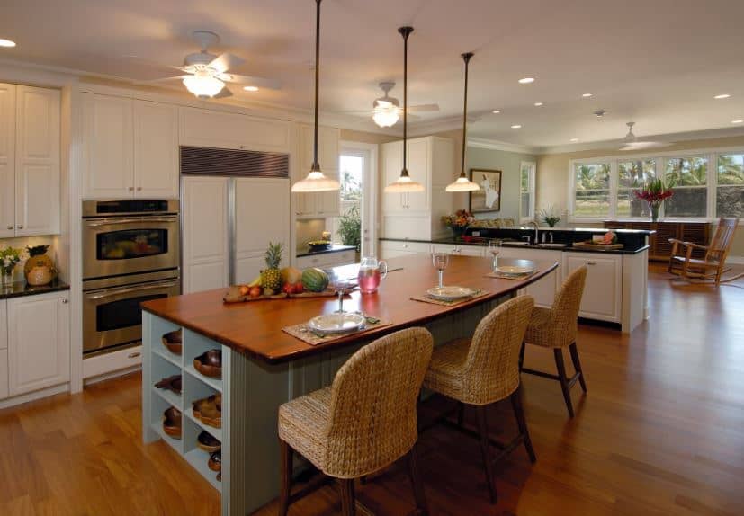 The woven wicker chairs complement the light blue wooden kitchen island as well as its wooden countertop with a built-in breakfast bar. These are all warmed by the various sources of warm yellow lights like the pendant lights and the ceiling fans.