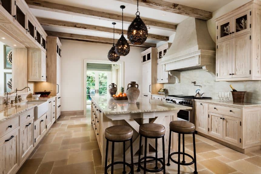 The charming decorative pendant lights stand out against the light wooden hues of the exposed wooden beams of the white ceiling. These beams match the wooden cabinetry of the two peninsulas and the large kitchen island in the middle.