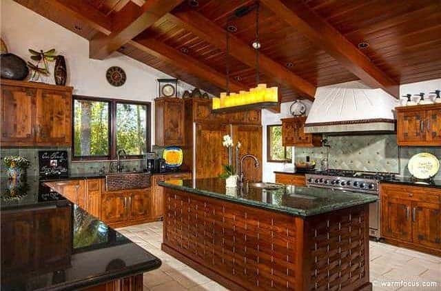 The wooden shed ceiling has exposed wooden beams that contrast the white upper walls. These white upper walls are adorned with various decors and dishware placed on the tops of the wooden floating cabinets with the same hue as the peninsula and its kitchen island.