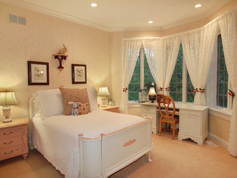 Small girl's bedroom with white classy walls and window curtains. The desk is placed near the windows overlooking the beautiful outdoor area surrounding the house.