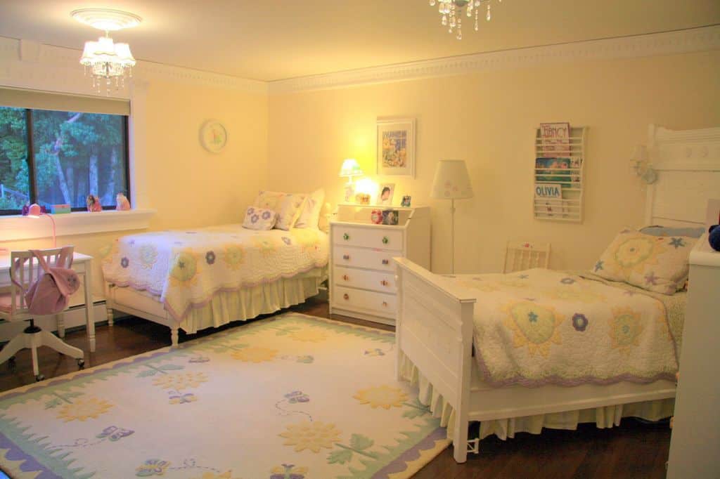 This girls bedroom offers two beds and a cute rug set on the hardwood flooring. The warm white lights look perfect together with the white walls.