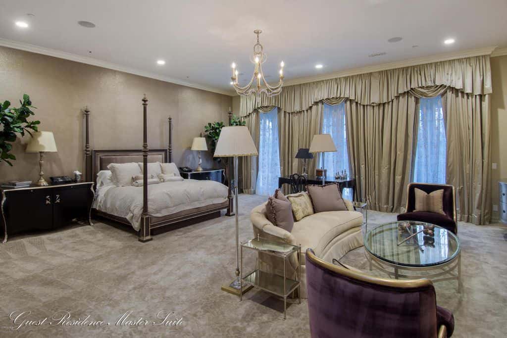 Luxury primary bedroom with velvet carpet flooring and full height windows dressed in classy drapes and valances. It includes a four-poster bed and a seating area illuminated by a candle chandelier and recessed ceiling lights.