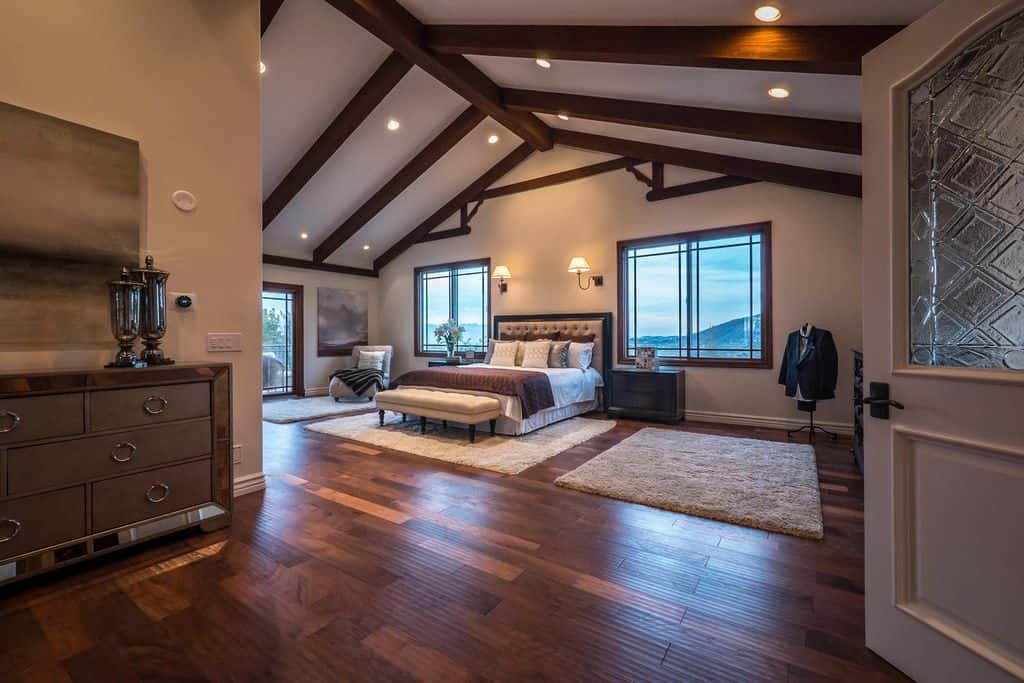 Recessed ceiling lights and wall sconces illuminate this spacious primary bedroom showcasing a tufted bed and a chaise lounge over gray shaggy rugs. It has hardwood flooring and a cathedral ceiling lined with dark wood beams.