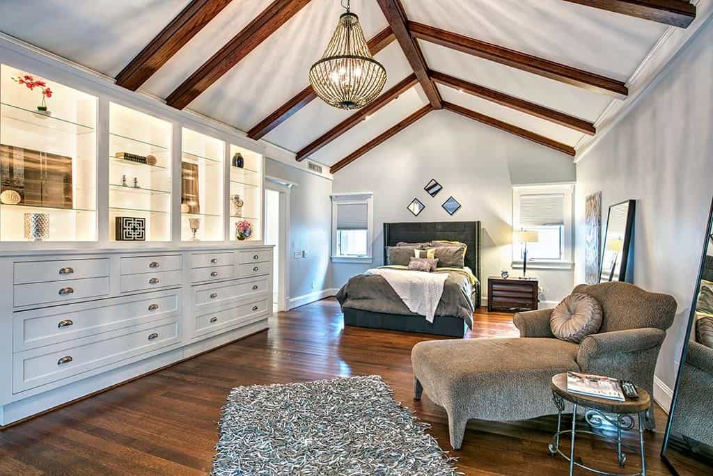 A classy chaise lounge flanked by full-length mirrors faces the large built-in cabinet that's fixed against the light gray wall. It is accompanied by a wingback bed and a warm chandelier that hung from the cathedral ceiling lined with wood beams.