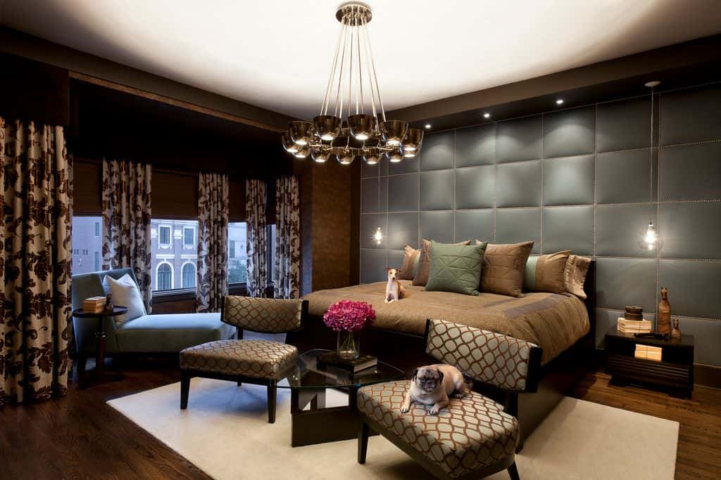 A tufted accent wall sets a cozy backdrop to the dark wood bed lighted by glass pendants and a contemporary chandelier. There's a seating area on its end and a comfy chaise lounge by the glazed windows dressed in floral draperies.