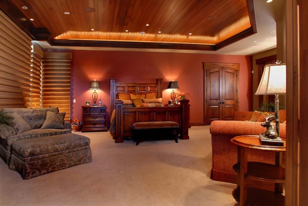 Warm master bedroom furnished with cozy seats and a wooden bed matching with the nightstands that are topped with stylish table lamps. It has carpet flooring and a tray ceiling clad in dark wood planks.