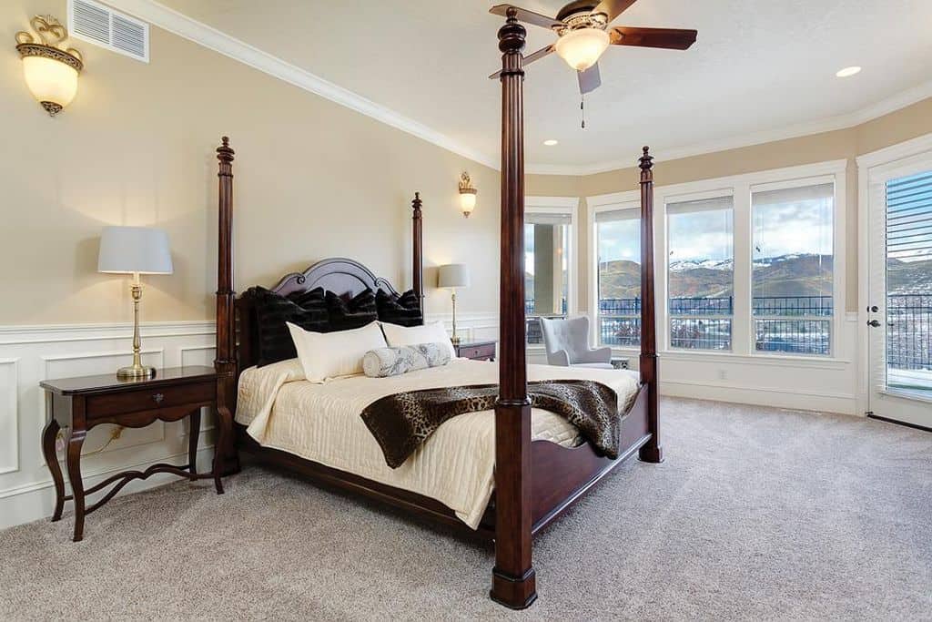 Sophisticated primary bedroom with gray carpet flooring and glass paneled windows overlooking a stunning mountain view. It includes a tufted wingback chair and a four-poster bed flanked by matching nightstands and brass table lamps.