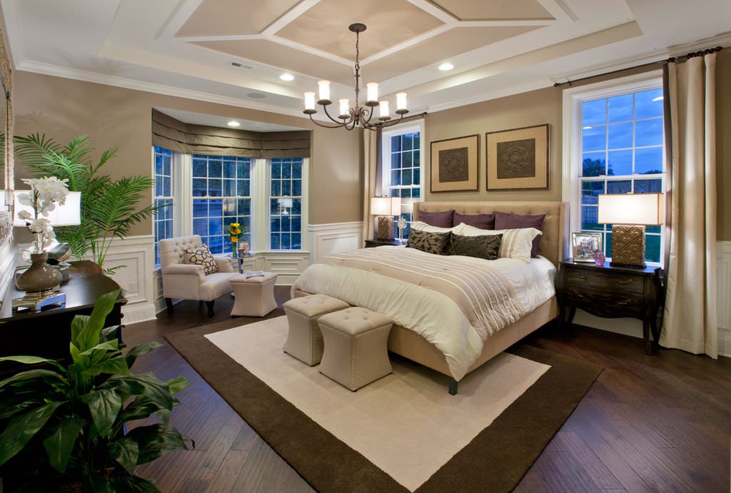 Beige tufted stools sit in front of the wingback bed facing the dark wood cabinet that matches the nightstands. There's a cozy chair by the bay window covered in taupe roman shades.